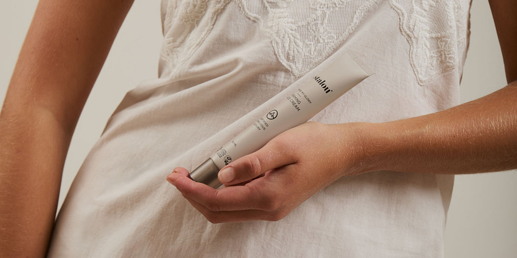 Close-up of a person holding a tube of Astalou Firming Face Cream against a white embroidered top in a studio setting
